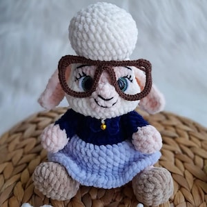 May include: A handmade crochet sheep doll wearing glasses, a blue dress, and a white hat. The sheep has pink ears and a small gold bell on its collar. The doll is sitting on a woven basket.