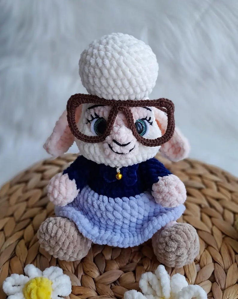 May include: A handmade crochet sheep doll wearing glasses, a blue dress, and a white hat. The sheep has pink ears and a small gold bell on its collar. The doll is sitting on a woven basket.