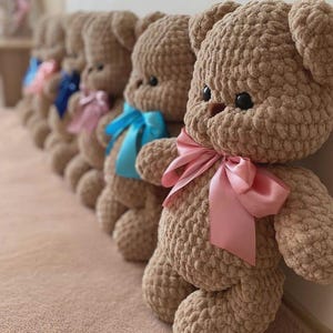 May include: A row of five handmade crochet teddy bears made from soft, light brown plush yarn. Each bear features dark round eyes, a small brown nose, and a satin ribbon tied around its neck in various colors including pink, blue, and light pink.