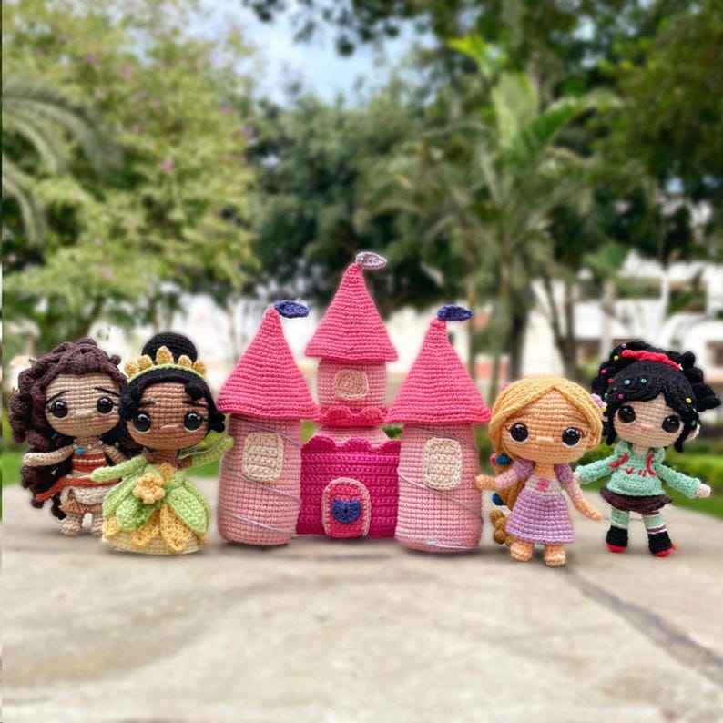 May include: A selection of handmade crochet dolls and a castle. The dolls feature characters with diverse skin tones, dressed in vibrant outfits. The castle is pink with a dark pink door and blue details. The dolls are positioned on a concrete surface.