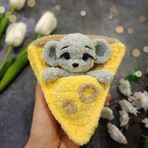 Puede incluir: Un juguete de ratón de ganchillo hecho a mano con cuerpo gris, ojos azules y largas pestañas, metido en una cuña de queso amarilla y marrón. El queso tiene detalles circulares marrones. El juguete se sostiene en una mano.