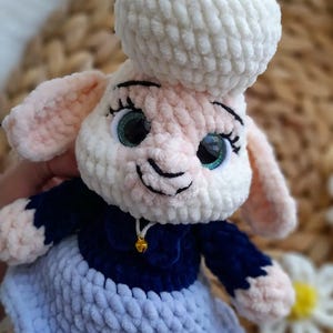 May include: A crocheted sheep doll with a white head and body, pink ears, and green eyes. It is dressed in a navy blue top and a light blue skirt. A small gold bell hangs from its neck. The doll is handmade.