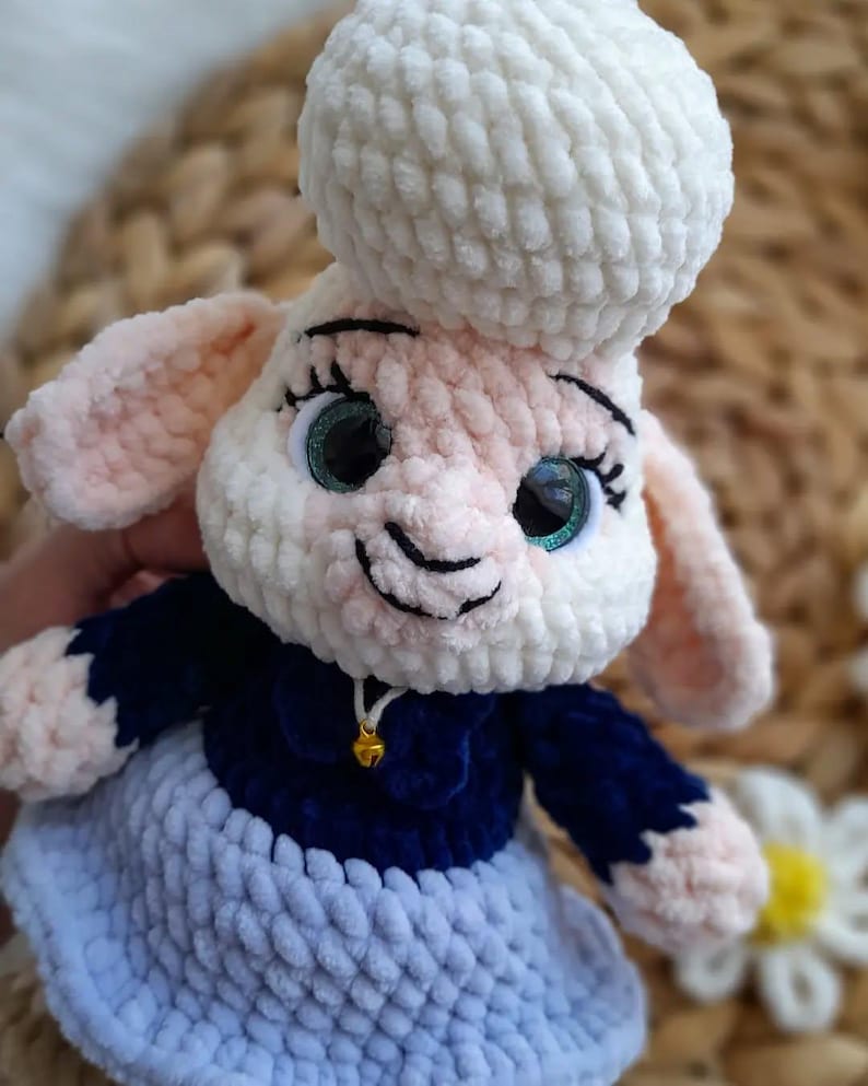 May include: A crocheted sheep doll with a white head and body, pink ears, and green eyes. It is dressed in a navy blue top and a light blue skirt. A small gold bell hangs from its neck. The doll is handmade.