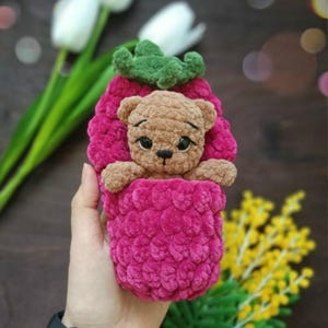 Peut inclure: Un étui en forme de framboise crocheté à la main, rose vif, avec un dessus en forme de feuille verte. Un petit ours en peluche beige sort de l'étui. L'étui est tenu dans une main, avec des fleurs en arrière-plan.