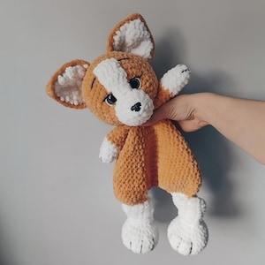 May include: A handmade plush dog toy in shades of orange, white, and brown. The dog has large, floppy ears, a white snout, and white paws. The toy is made of a soft, textured material.