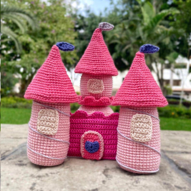 May include: A handcrafted crochet castle in various shades of pink. The castle has three towers with conical roofs, small windows, and decorative details. The central tower is a darker pink, and the other two are a lighter pink. A whimsical decorative item.