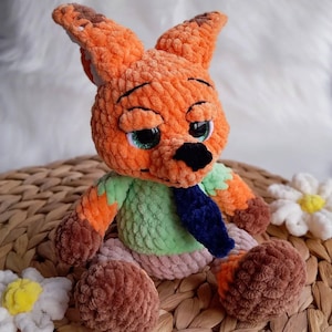 May include: A handmade orange and brown crochet fox doll with a green shirt and blue tie. The plush toy has large, expressive eyes with green irises and a black nose. The fox is sitting on a woven surface, with white flower accents.