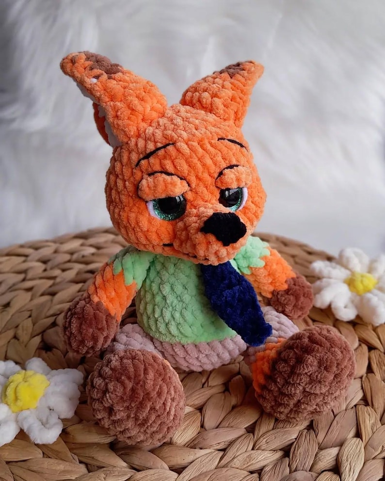 May include: A handmade orange and brown crochet fox doll with a green shirt and blue tie. The plush toy has large, expressive eyes with green irises and a black nose. The fox is sitting on a woven surface, with white flower accents.