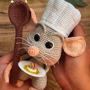 Peut inclure: Une poupée souris crochetée portant une toque de chef blanche, tenant une cuillère en bois crochetée marron dans une main et une petite assiette blanche avec un aliment miniature coloré dans l'autre. La souris a une fourrure grise, des yeux noirs.