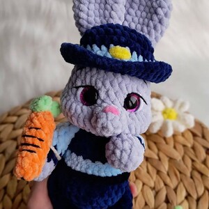 May include: A handmade crochet bunny doll in shades of lavender, navy, and orange. The bunny wears a navy blue hat with a yellow accent and holds an orange carrot. The doll has large, sparkly eyes and a pink nose.