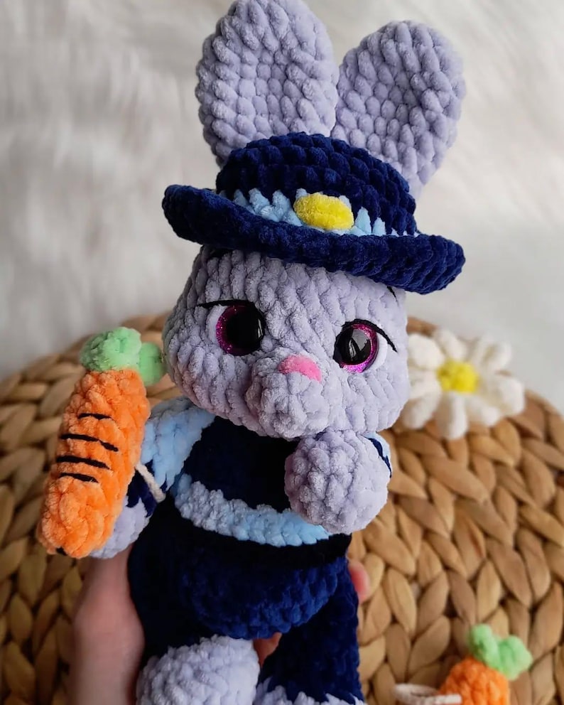 May include: A handmade crochet bunny doll in shades of lavender, navy, and orange. The bunny wears a navy blue hat with a yellow accent and holds an orange carrot. The doll has large, sparkly eyes and a pink nose.