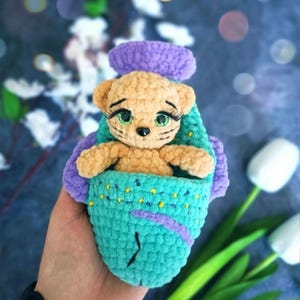 Puede incluir: Un juguete de ganchillo hecho a mano con un osito de peluche de color canela con ojos verdes y largas pestañas, metido en un portabebés turquesa y morado. El portabebés tiene un aspecto texturizado y cosido, con detalles morados y puntos amarillos.