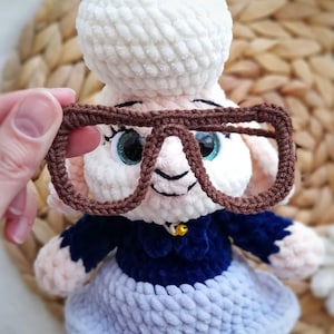May include: A handmade crochet sheep doll with a white head, a blue dress, and brown glasses. The doll has blue eyes and a small gold bell on its collar. The doll is made of soft, textured yarn.