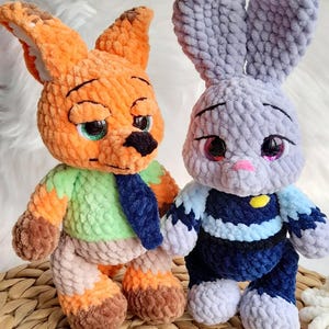 May include: Two handmade crochet stuffed animals: an orange fox with green and blue accents and a gray bunny with blue and white accents. Both have detailed facial features and are made with soft, textured yarn. The fox has green eyes, and the bunny has pink eyes.