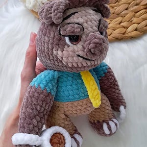 May include: A handmade crochet plush toy in the shape of a cartoon character. The plushie has a brown and tan body, a blue shirt, and a yellow tie. The toy has white paws and a smiling face.