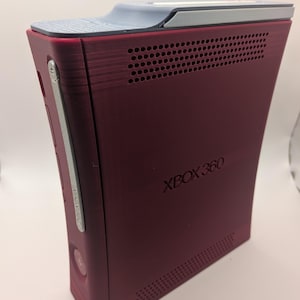 May include: A burgundy Xbox 360 console with a gray top and silver accents. The console has the text "XBOX 360" printed on the side. The console has a series of ventilation holes.