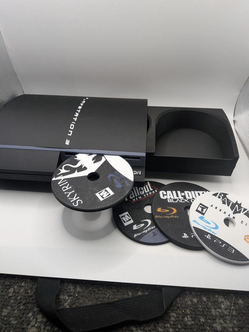 May include: A black PlayStation 3 console with several game discs. The discs include "Skyrim", "Fallout: New Vegas", "Call of Duty: Black Ops", and "Batman: Arkham City". The console is open, with one disc partially inserted.