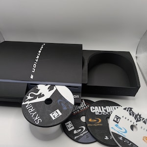 May include: A black PlayStation 3 console with a disc tray open, displaying the game disc "Skyrim." Other game discs include "Fallout: New Vegas," "Call of Duty: Black Ops," and "Batman: Arkham City." The console is on a white surface.