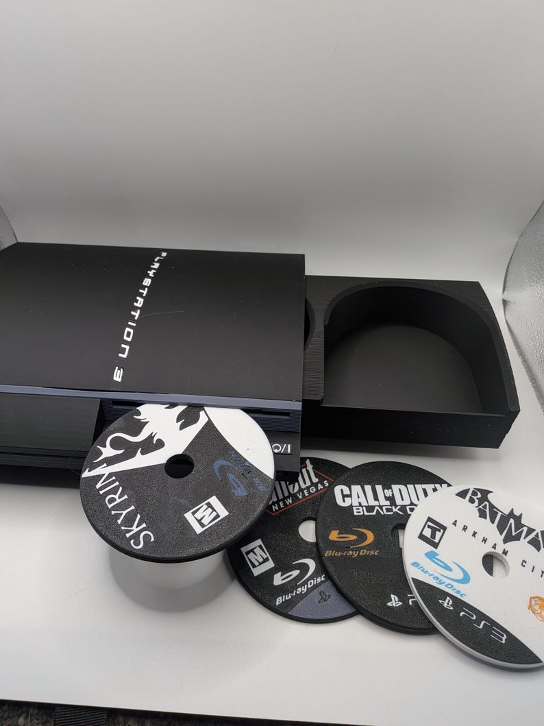 May include: A black PlayStation 3 console with a disc tray open, displaying the game disc "Skyrim." Other game discs include "Fallout: New Vegas," "Call of Duty: Black Ops," and "Batman: Arkham City." The console is on a white surface.