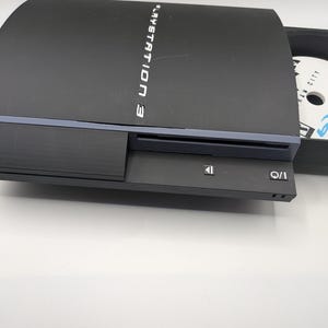 May include: Black PlayStation 3 console with a disc tray open, revealing a game disc. The console has the words "PlayStation 3" printed on the top. The I/O and eject buttons are visible on the front.