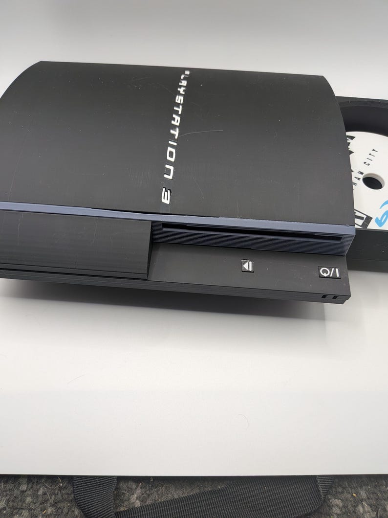 May include: Black PlayStation 3 console with a disc tray open, revealing a game disc. The console has the words "PlayStation 3" printed on the top. The I/O and eject buttons are visible on the front.