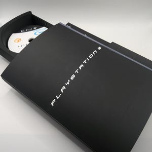 May include: Black PlayStation 3 console with the disc tray partially open, revealing a game disc. The words "PLAYSTATION 3" are printed in white on the console's cover. The disc has a blue and white design.