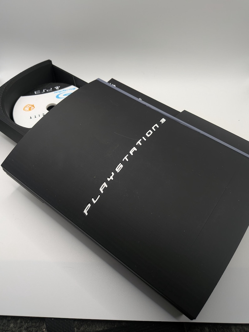 May include: Black PlayStation 3 console with the disc tray partially open, revealing a game disc. The words "PLAYSTATION 3" are printed in white on the console's cover. The disc has a blue and white design.