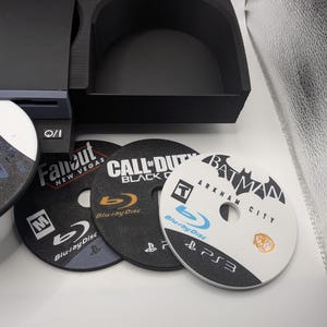 May include: A black console-style organizer with three game discs. The discs are labeled "Fallout New Vegas", "Call of Duty Black Ops", and "Batman Arkham City". The discs are Blu-ray.