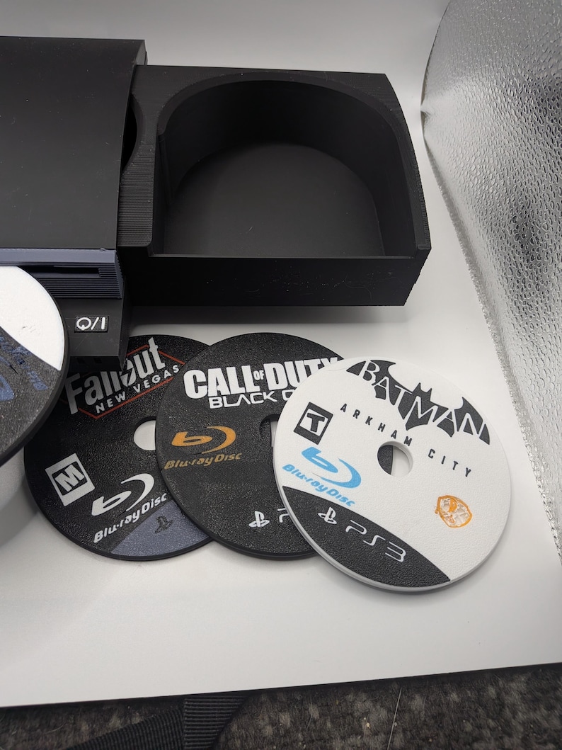 May include: A black console-style organizer with three game discs. The discs are labeled "Fallout New Vegas", "Call of Duty Black Ops", and "Batman Arkham City". The discs are Blu-ray.