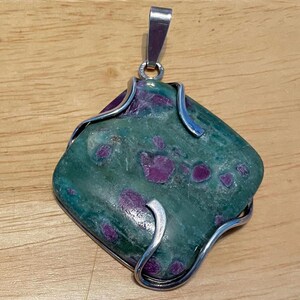 May include: A square-shaped pendant featuring a polished green stone with embedded magenta-colored inclusions. The stone is secured by a silver-toned metal wire frame that wraps around the edges, and it includes a silver-toned metal bail at the top.