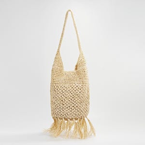 May include: A woven straw tote bag with a long shoulder strap. The bag is beige with a fringe detail at the bottom. The bag is made of natural materials and is suitable for carrying items.