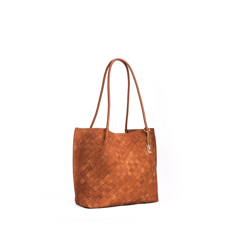 May include: A brown woven leather tote bag with long handles. The bag has a rectangular shape and a woven pattern. A small gold-colored charm hangs from one of the handles. The bag is set against a white background.