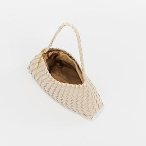 May include: A cream-colored woven handbag with a braided handle. The bag has a rectangular shape with a rounded bottom and a gold-colored clasp. The interior of the bag is a light brown color.