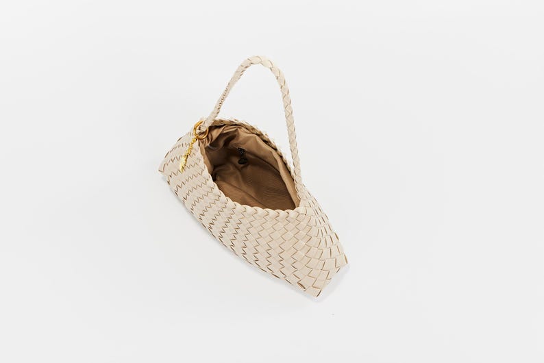 May include: A cream-colored woven handbag with a braided handle. The bag has a rectangular shape with a rounded bottom and a gold-colored clasp. The interior of the bag is a light brown color.
