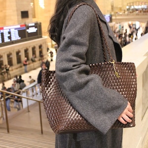Pode incluir: Uma bolsa tote marrom tecida com uma al&ccedil;a tran&ccedil;ada e detalhes de corrente dourada. A bolsa &eacute; segurada por uma pessoa que usa um casaco cinza. O fundo mostra uma esta&ccedil;&atilde;o ferrovi&aacute;ria movimentada com pessoas e escadas.