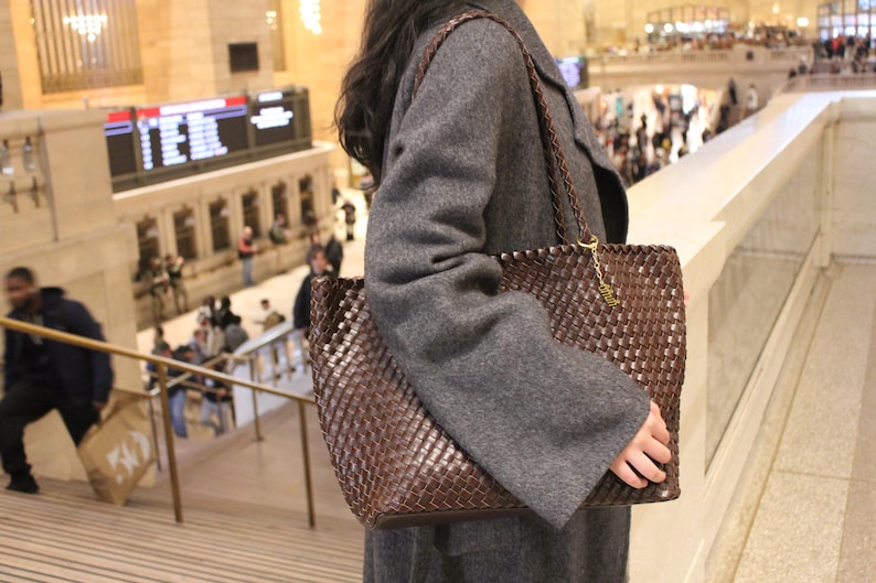 Pode incluir: Uma bolsa tote marrom tecida com uma al&ccedil;a tran&ccedil;ada e detalhes de corrente dourada. A bolsa &eacute; segurada por uma pessoa que usa um casaco cinza. O fundo mostra uma esta&ccedil;&atilde;o ferrovi&aacute;ria movimentada com pessoas e escadas.