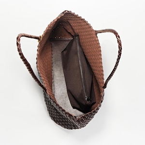 Pode incluir: Uma bolsa tote marrom tecida com al&ccedil;as tran&ccedil;adas. A bolsa tem formato retangular e parte superior aberta, revelando um interior marrom escuro com um bolso com z&iacute;per. A bolsa &eacute; feita de um material texturizado.