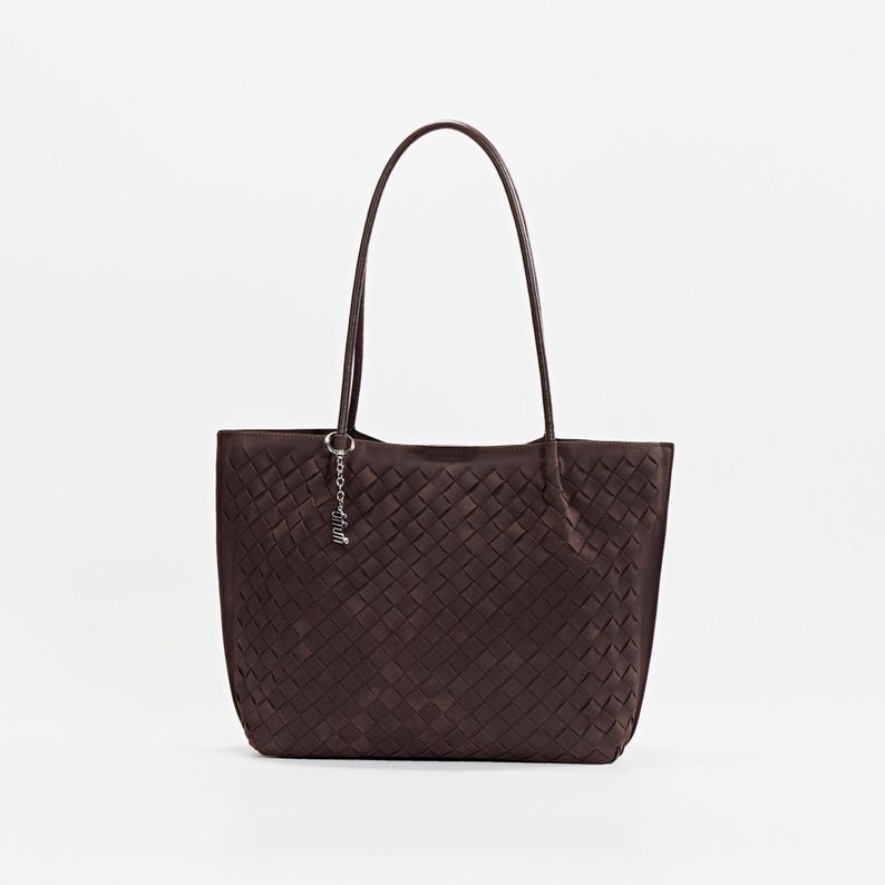 May include: A dark brown tote bag with a woven pattern. The bag has two long handles and a small silver charm attached to one side. The bag is made of a soft material and is suitable for everyday use.