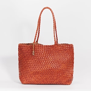 Pode incluir: Uma bolsa tote de couro tecido em um tom marrom avermelhado. Apresenta al&ccedil;as tran&ccedil;adas e um pequeno pingente dourado. O design da bolsa apresenta um padr&atilde;o de tecido de cesta, oferecendo um acess&oacute;rio elegante e espa&ccedil;oso.
