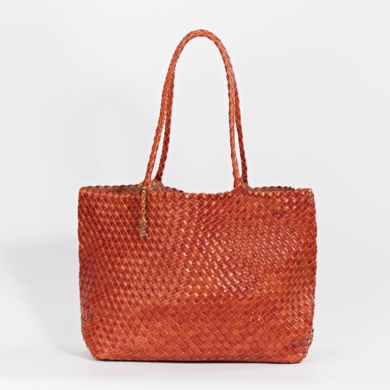Pode incluir: Uma bolsa tote de couro tecido em um tom marrom avermelhado. Apresenta al&ccedil;as tran&ccedil;adas e um pequeno pingente dourado. O design da bolsa apresenta um padr&atilde;o de tecido de cesta, oferecendo um acess&oacute;rio elegante e espa&ccedil;oso.