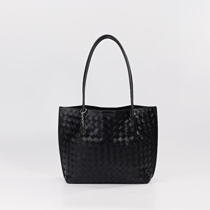 May include: Black tote bag with a woven design. The bag has two long handles and a small charm attached to the interior. The bag is made of a shiny material and is set against a white background.