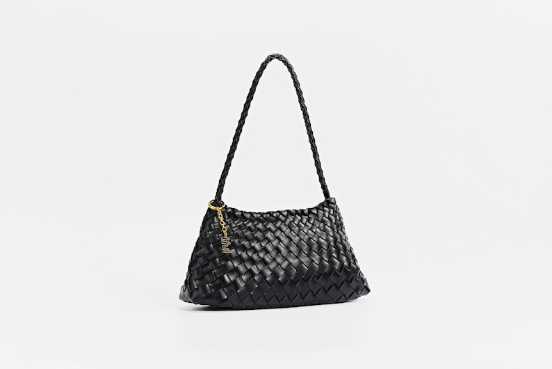 May include: Black woven shoulder bag with a braided handle. The bag features a gold-colored charm and a rectangular shape. The bag is made of a woven material and has a braided handle. The background is plain white.