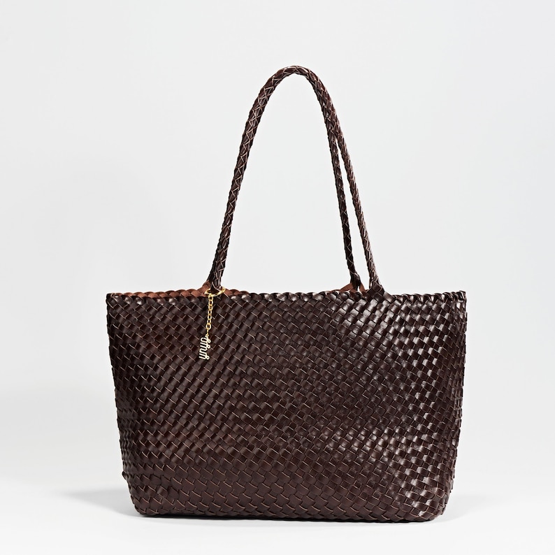 Pode incluir: Uma bolsa tote tecida marrom escuro com al&ccedil;as tran&ccedil;adas. A bolsa tem formato retangular e um pingente dourado pendurado na al&ccedil;a. A bolsa &eacute; feita de um material tecido, e as al&ccedil;as tamb&eacute;m s&atilde;o tran&ccedil;adas.