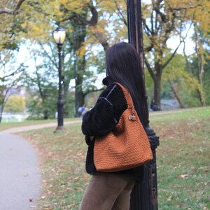 K&ouml;nnte beinhalten: Eine strukturierte, orangefarbene Tragetasche mit Schulterriemen. Die Tasche wird von einer Person getragen, die einen schwarzen Mantel und eine braune Hose tr&auml;gt. Der Hintergrund zeigt einen Park mit B&auml;umen und einer Stra&szlig;enlaterne.