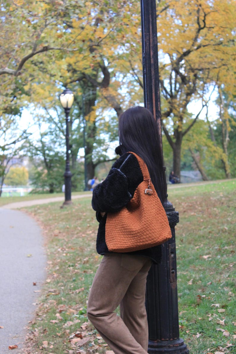 K&ouml;nnte beinhalten: Eine strukturierte, orangefarbene Tragetasche mit Schulterriemen. Die Tasche wird von einer Person getragen, die einen schwarzen Mantel und eine braune Hose tr&auml;gt. Der Hintergrund zeigt einen Park mit B&auml;umen und einer Stra&szlig;enlaterne.