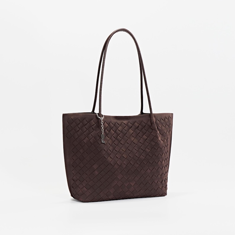 May include: A brown tote bag with a woven pattern. The bag has two long handles and a small charm hanging from one handle. The bag is made of a soft material and has a rectangular shape.
