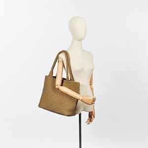 May include: A tan tote bag with a woven texture is displayed on a mannequin. The bag has two braided handles and a rectangular shape. The mannequin is positioned against a white background, showcasing the bag's design and color.