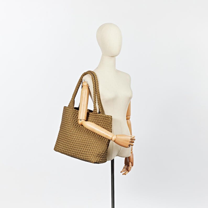 May include: A tan tote bag with a woven texture is displayed on a mannequin. The bag has two braided handles and a rectangular shape. The mannequin is positioned against a white background, showcasing the bag's design and color.