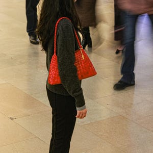 May include: A red woven handbag with a shoulder strap is carried by a person wearing a dark gray sweater and black pants. The bag has a gold-colored chain and clasp. The background shows blurred figures walking on a light-colored tiled floor.