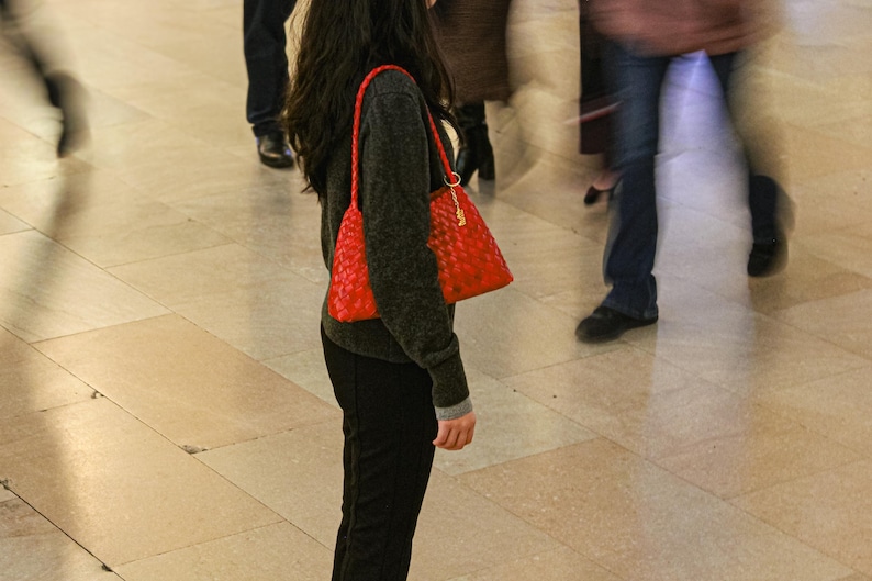 May include: A red woven handbag with a shoulder strap is carried by a person wearing a dark gray sweater and black pants. The bag has a gold-colored chain and clasp. The background shows blurred figures walking on a light-colored tiled floor.